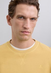 Junger Mann mit kurzen lockigen braunen Haaren und blauen Augen, der einen gelben Rundhalsausschnitt-Pullover über einem weißen Hemd trägt, neutraler Hintergrund.