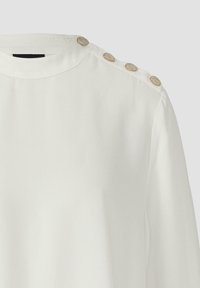 Weiße Bluse mit rundem Ausschnitt, verziert mit goldfarbenen Knöpfen an der Schulter. Glatter, leichter Stoff mit leichtem Glanz.