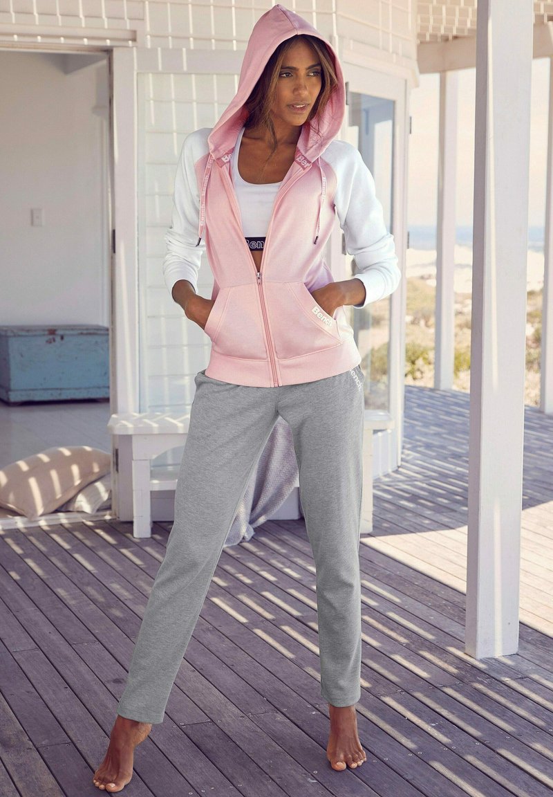 Helle rosa Zip-up Hoodie mit weißen Ärmeln, ausgestattet mit einer Kapuze und Taschen, kombiniert mit grauen Sweatpants. Beide Kleidungsstücke haben eine entspannte Passform.