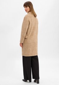 Cappotto beige in misto lana con un motivo a quadri, maniche lunghe e tasche laterali, abbinato a pantaloni neri a gamba larga e scarpe a punta.