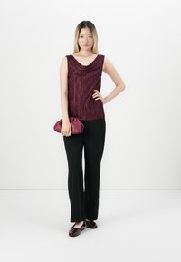 Jongem vrouw staat tegen een witte achtergrond, gekleed in een mouwloze plooien burgundy top, zwarte broek, burgundy loafers en houdt een bijpassende clutch vast.