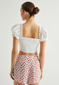 Weißes Crop-Top mit Puffärmeln, Augenlederdetails und gewellten Rändern; kombiniert mit hochtaillierten Shorts mit einem roten Blumenmuster.