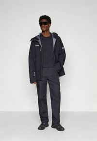 Crna jakna za vanjsku upotrebu s kapuljačom, sivim detaljima, odgovarajuće hlače, siva košulja ispod i sunčane naočale. Ima džepove na patentni zatvarač i vodootporan materijal.