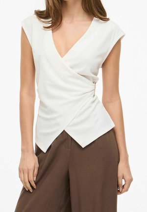 Femme portant un haut blanc sans manches croisé avec un détail épingle sur le côté et un pantalon large marron sur fond blanc.
