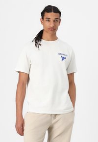 Camiseta de algodón color crema claro con el logo azul de "WRANGLER" y un gráfico de un toro en el lado izquierdo del pecho, manga corta y un corte relajado.