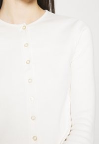 White, fitted cardigan with a round neckline, seven button closure featuring pearl-like buttons, and long sleeves made of soft material.