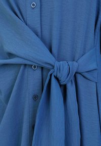Blue button-up garment featuring a tied waist detail, made from lightweight, textured fabric with a smooth finish.