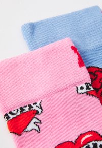 Deux paires de chaussettes montantes : une rose avec des motifs de cœurs rouges et une bleue claire. Les deux ont une texture côtelée et des poignets de couleur unie.