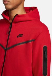 Röd Nike-hoodie med dragkedja framtill, svarta detaljer och en dragkedjefick på sidan. Tillverkad av ett mjukt, texturerat tyg.