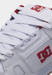Baskets blanches avec lacets blancs, logo DC rouge sur le côté et la languette, comprenant des détails perforés et des coutures visibles.