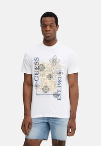 Camiseta blanca de algodón con diseños gráficos florales en negro y naranja que incluyen el texto "GUESS" y "EST. 1981", con cuello redondo y mangas cortas.