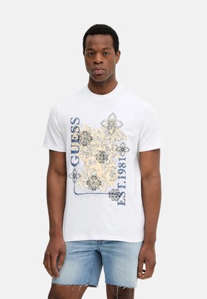 Camiseta blanca de algodón con diseños gráficos florales en negro y naranja que incluyen el texto "GUESS" y "EST. 1981", con cuello redondo y mangas cortas.