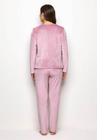 Donna in piedi a piedi nudi, girata di spalle, che indossa una morbida maglietta a maniche lunghe rosa pallido e pantaloni abbinati su uno sfondo bianco semplice.