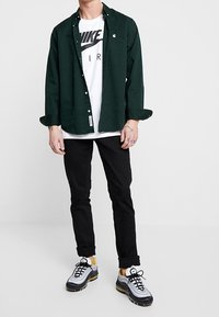 Dark green button-up shirt over a white T-shirt with a black logo, black pants, and gray sneakers with yellow accents.