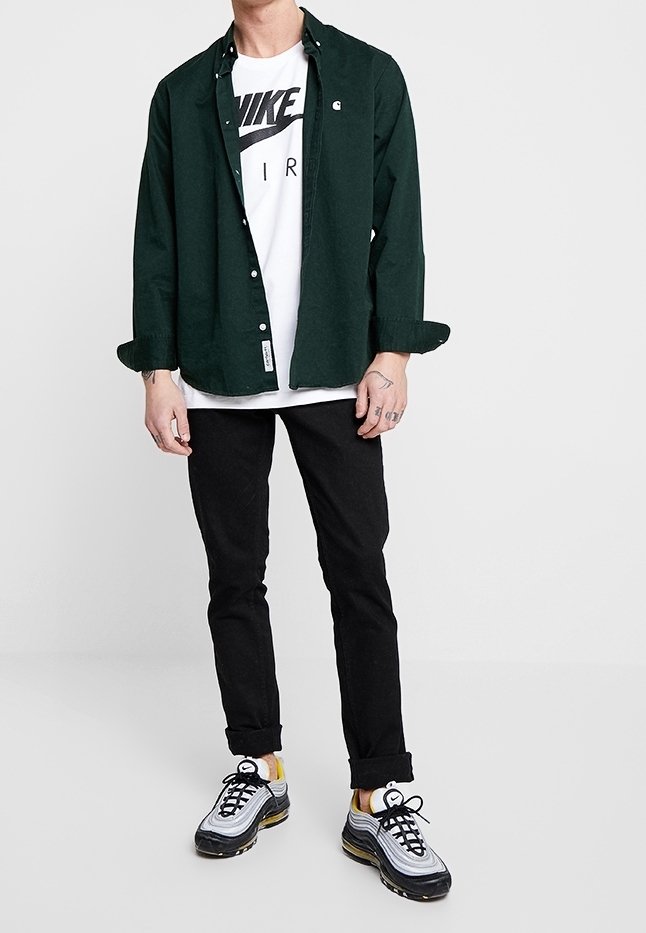 Dark green button-up shirt over a white T-shirt with a black logo, black pants, and gray sneakers with yellow accents.