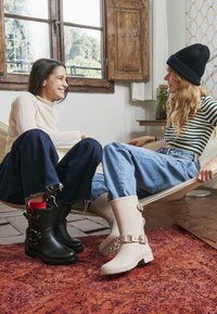 Dwie kobiety uśmiechają się, siedząc na hamaku w środku, ubrane w casualowe ubrania i kontrastujące czarne i beżowe buty na czerwonym dywanie.