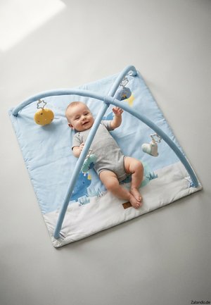 Baby lying on blue play mat with soft hanging toys, smiling and reaching out while wearing a gray romper.