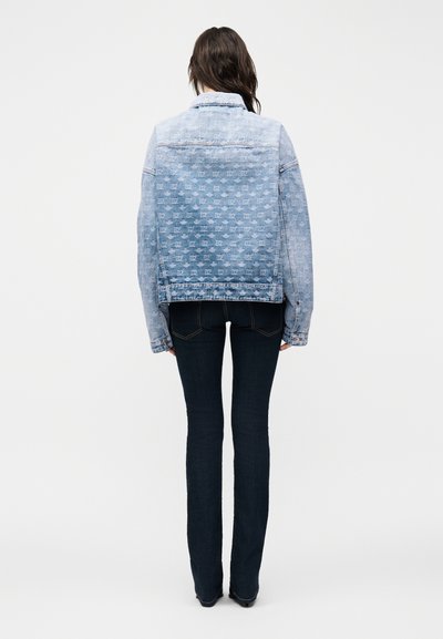 Femme portant une veste en jean bleu clair à motifs et un jean slim bleu foncé, debout de dos sur un fond uni.