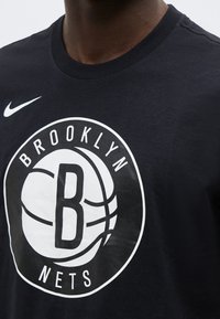 Κοντινό πλάνο προσώπου που φοράει μαύρο μπλουζάκι με λευκό λογότυπο των Brooklyn Nets και το σήμα Nike στη μέση.