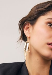 Guldgeometriska örhängen med hängande rektangulära former som skapar en kaskadeffekt. Slät metallisk yta, fästa med en tunn stift.