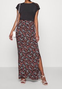 Svart topp med spetsdetaljer; blommig maxikjol i svart, röd och vit med sid slits; vita sandaler med remmar; liten vit axelväska.