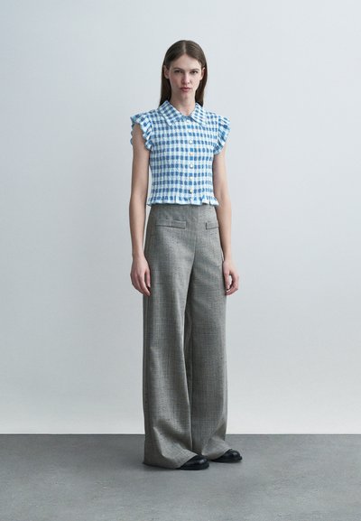 Γυναίκα όρθια που φοράει μπλε και λευκό καρό γυναικείο αμάνικο μπλουζάκι και γκρι φαρδιά παντελόνια ψηλόμεσα με μαύρα παπούτσια.