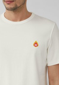 Weißes Baumwoll-T-Shirt mit Rundhalsausschnitt, das auf der linken Brust eine kleine gestickte Flammengrafik mit "BBQ" in Rot und Gelb zeigt.