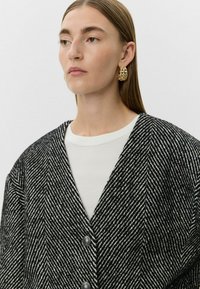 Oversized blazer s houndstooth vzorem v černobílé barvě, s véčkovým výstřihem, dvěma tmavými knoflíky a texturovanou tkaninou. Pod ním bílý kulatý výstřih.