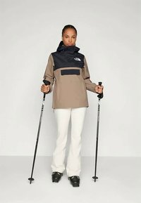 Időjárásálló dzseki, barna testtel és sötétkék felsőrésszel, elöl zsebbel, fehér nadrággal és fekete sícipővel párosítva.