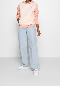 Person som bär en rosa och persiko Nike-hoodie, ljusblå vida jeans och vita sneakers, stående mot en enkel vit bakgrund.