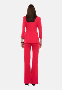 Completo su misura rosso con giacca aderente e pantaloni svasati. Tessuto liscio, maniche a tre quarti e una silhouette pulita e moderna.