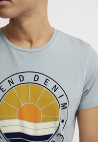 Lys blå crew neck t-skjorte med et rundt grafisk motiv av en solnedgang i oransje og gult, lagdelte bølger i krem og marineblått, og svart tekst.