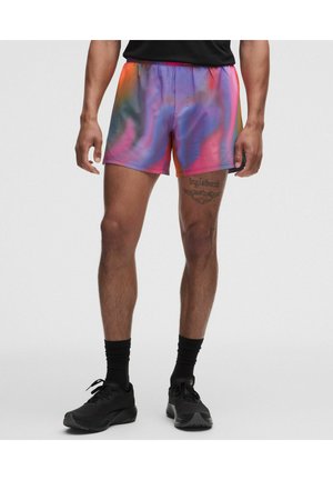 Hombre vistiendo pantalones deportivos teñidos en varios colores, calcetines negros y zapatillas negras, de pie con los pies ligeramente separados sobre un fondo liso.