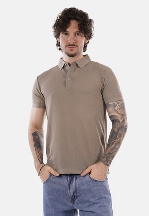 Uomo con capelli ricci e tatuaggi, che indossa una polo beige con trama e jeans blu, in piedi con le mani nelle tasche su sfondo bianco.