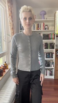Gray ribbed cardigan with a round neckline and button closure. Worn with black loose-fitting pants. Positioned in a bright indoor setting.