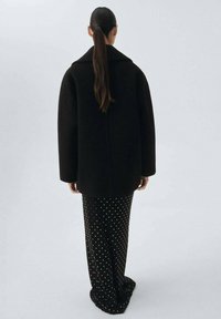 Persona con un lungo ponytail che indossa un cappotto oversize nero e una gonna nera con pois bianchi, in piedi di fronte a uno sfondo chiaro e semplice.