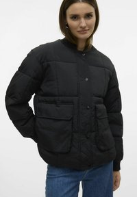 Veste noire matelassée avec des boutons-pression, un col montant et deux poches plaquées à l'avant. Fabriquée en tissu texturé avec une finition mate.