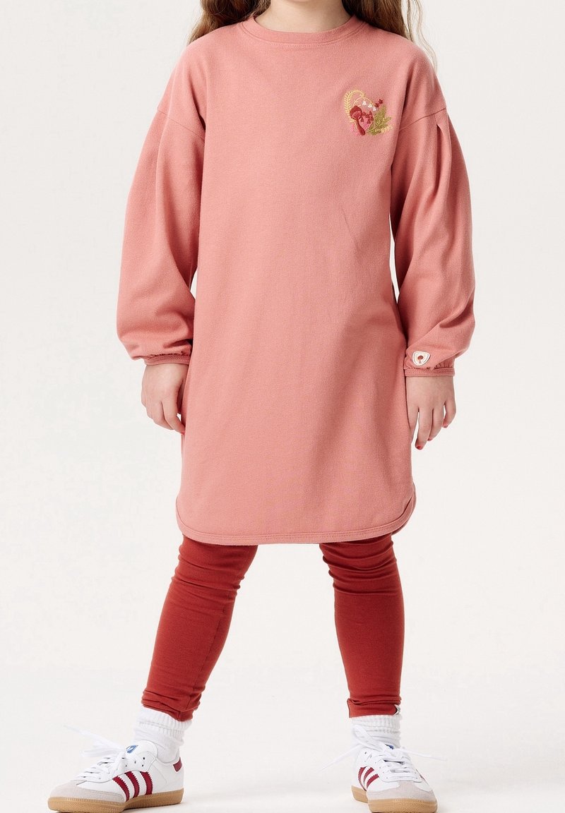 Enfant portant une robe rose à manches longues avec une broderie florale, associée à des leggings couleur rouille et des baskets blanches à rayures rouges.
