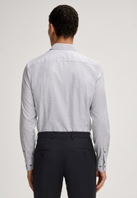 Helles graues Langarmshirt mit einem dezenten Muster; mit knöpfbaren Manschetten versehen. Getragen mit dunkel taillierten Hosen.