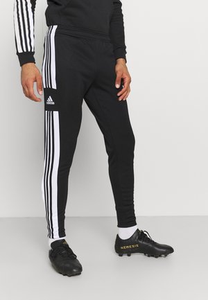 Osoba nosząca czarne spodnie dresowe Adidas z białymi paskami i logo, w zestawieniu z czarnymi korkami i białymi skarpetami, stojąca na szarej podłodze.