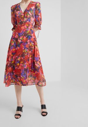 Nainen pukeutuneena polvipituiseen, kukkakuviolliseen mekkoon, jossa on liehukkeet hihnoissa, ja mustiin avokkaisiin kiilakorkoihin yksinkertaisella taustalla.