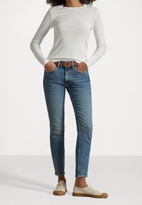 Vit långärmad topp, blå åtsittande jeans, brun läderbälte och ljusa sneakers. Enkel design och avslappnad stil.