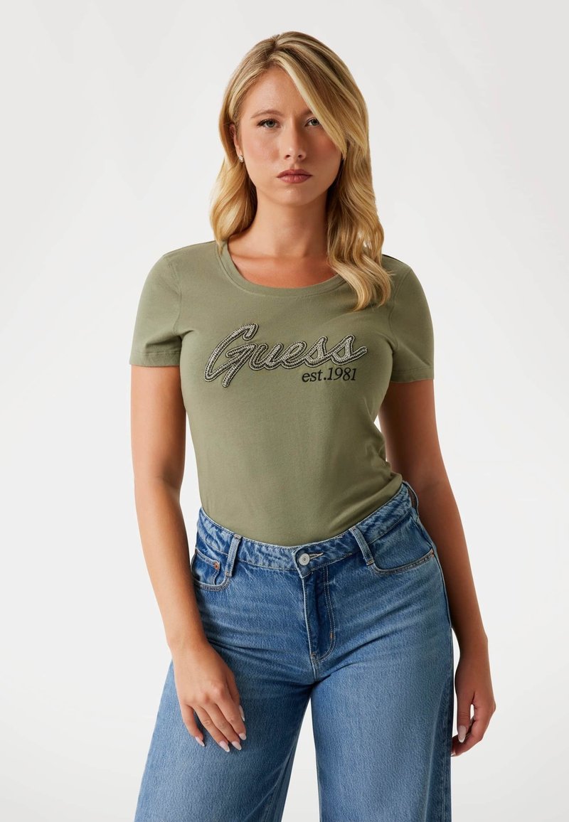 Olive green short-sleeve t-shirt with rounded neck, featuring "Guess est. 1981" embroidered in black and silver. Worn with high-waisted blue jeans.