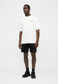 Mann steht vor weißem Hintergrund und trägt ein weißes übergroßes T-Shirt, schwarze Shorts, schwarze Socken und schwarze Turnschuhe.