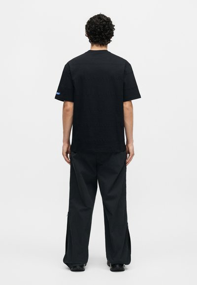 Άνδρας με σγουρά μαλλιά που φοράει απλό μαύρο t-shirt με κοντά μανίκια και φαρδιά μαύρα παντελόνια, στέκεται με την πλάτη σε λευκό φόντο.