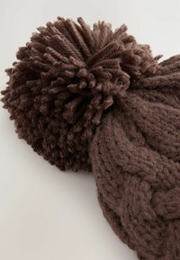 Brown knitted beanie with a large, textured pom-pom on top, featuring a cable knit pattern and a soft, fuzzy texture throughout.