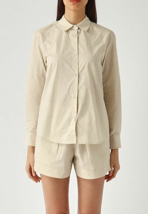 Donna che indossa una camicia beige a maniche lunghe con bottoni e pantaloncini coordinati, in piedi davanti a uno sfondo bianco neutro.