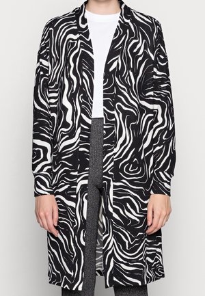 Schwarz-weiß gestreifter, zebra-musterter langer Cardigan, getragen über einem weißen Oberteil und dunkler, strukturierter Hose, die Hände entspannt an den Seiten.