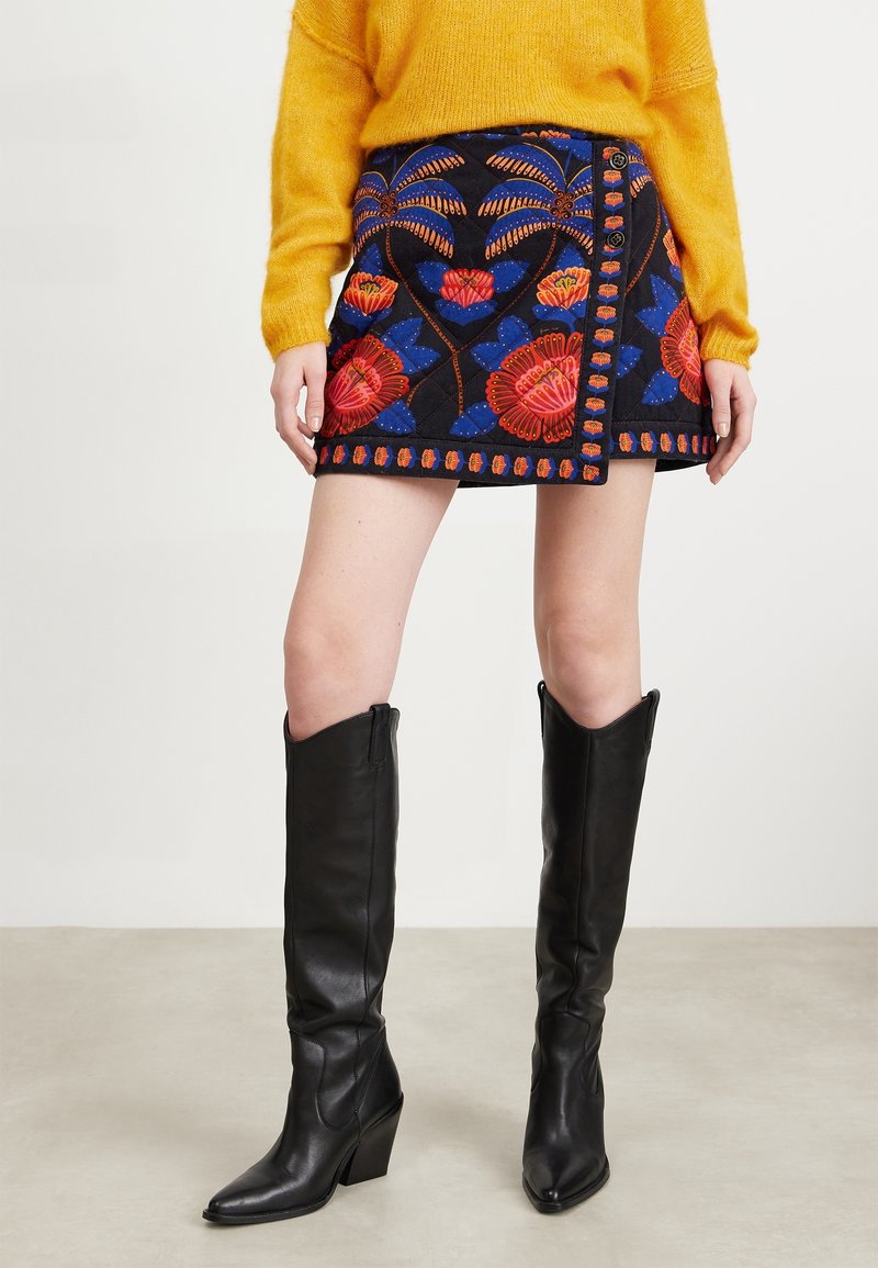 Personne portant un pull jaune, une mini-jupe noire à motifs floraux et des bottes noires à talons montant jusqu'aux genoux sur un fond uni.