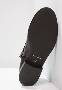 Bota negra con cremallera, inclinada hacia arriba, mostrando una suela lisa con tamaño 37 y el texto 'Hecho en Portugal'.